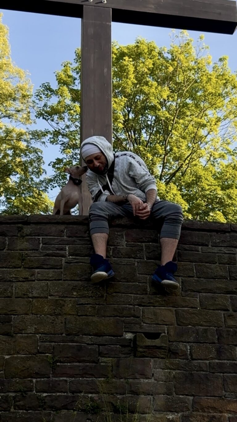 eazy sitzt in einem grauen Kapuzenpullover auf einer Steinmauer; azula steht neben ihm.