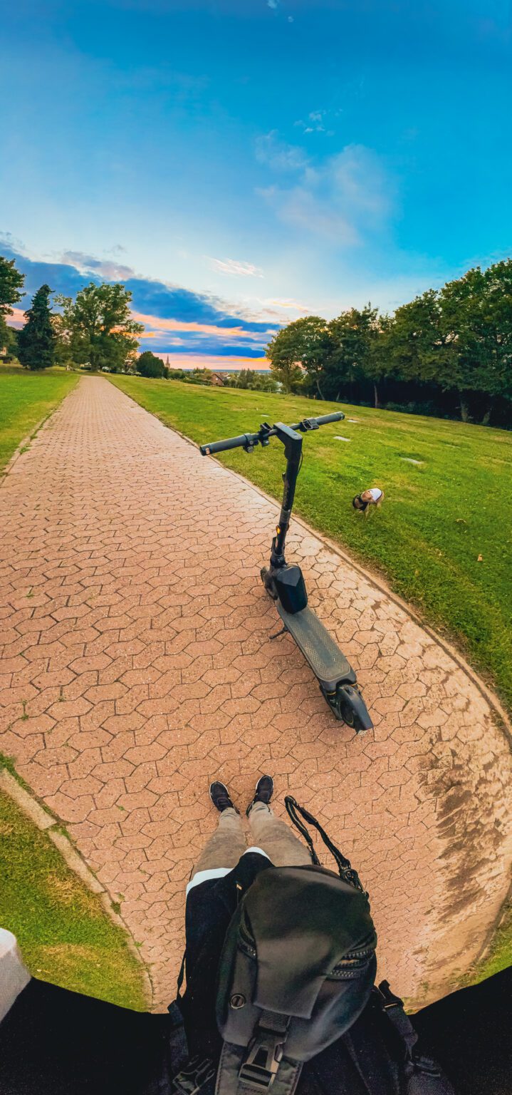 Blick über einen Rucksack und eazys Schuhe auf einen Roller am Parkweg; azula steht auf der angrenzenden Wiese.